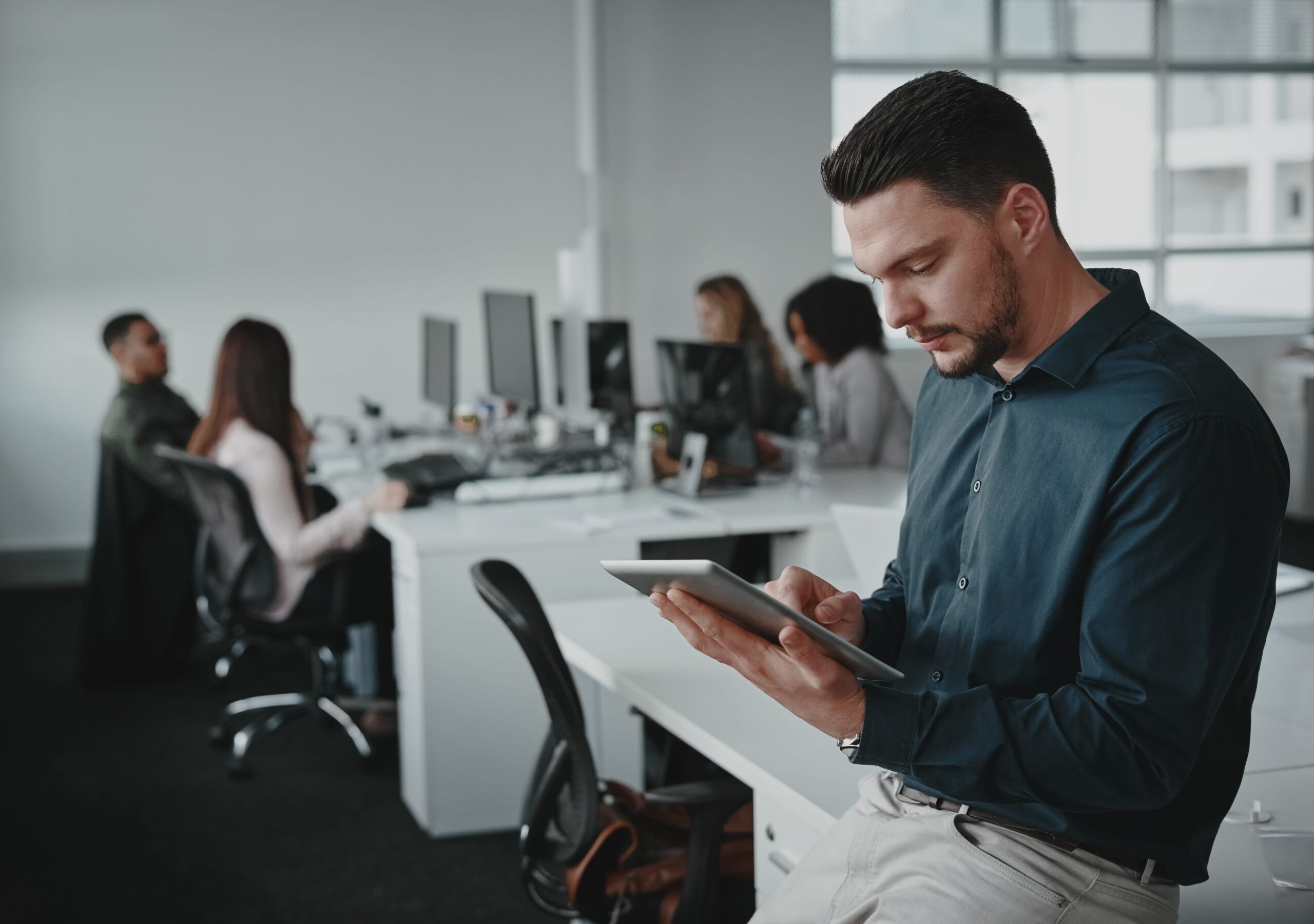 portrait young businessman leaning desk using tablet pc his colleagues working background 1 scaled